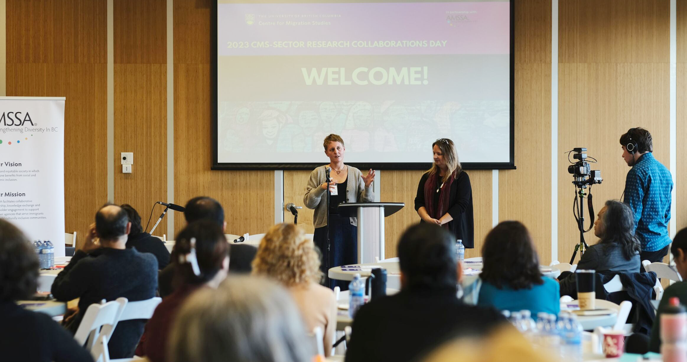 Antje Ellerman, Co-Director of UBC Centre for Migration Studies, and Katie Crocker, CEO of AMSSA, standing at a desk facing the audience during the 2023 Research Collaborations Day.